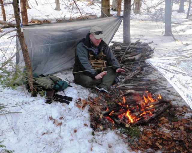 Man warming hands next to fire in snow while surrounded by a mylar blanket shelter