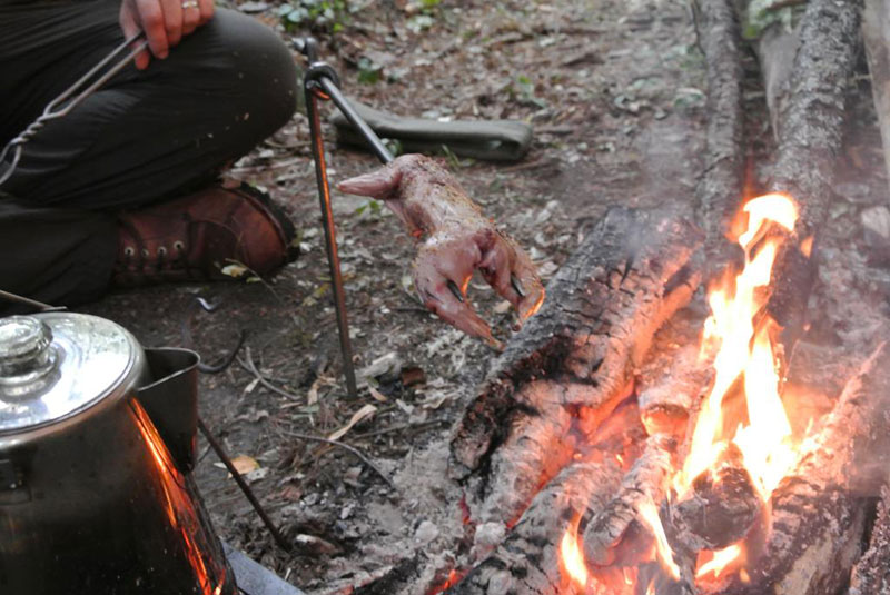 Human cooking a squirrel over a fire with a teapot and skewers